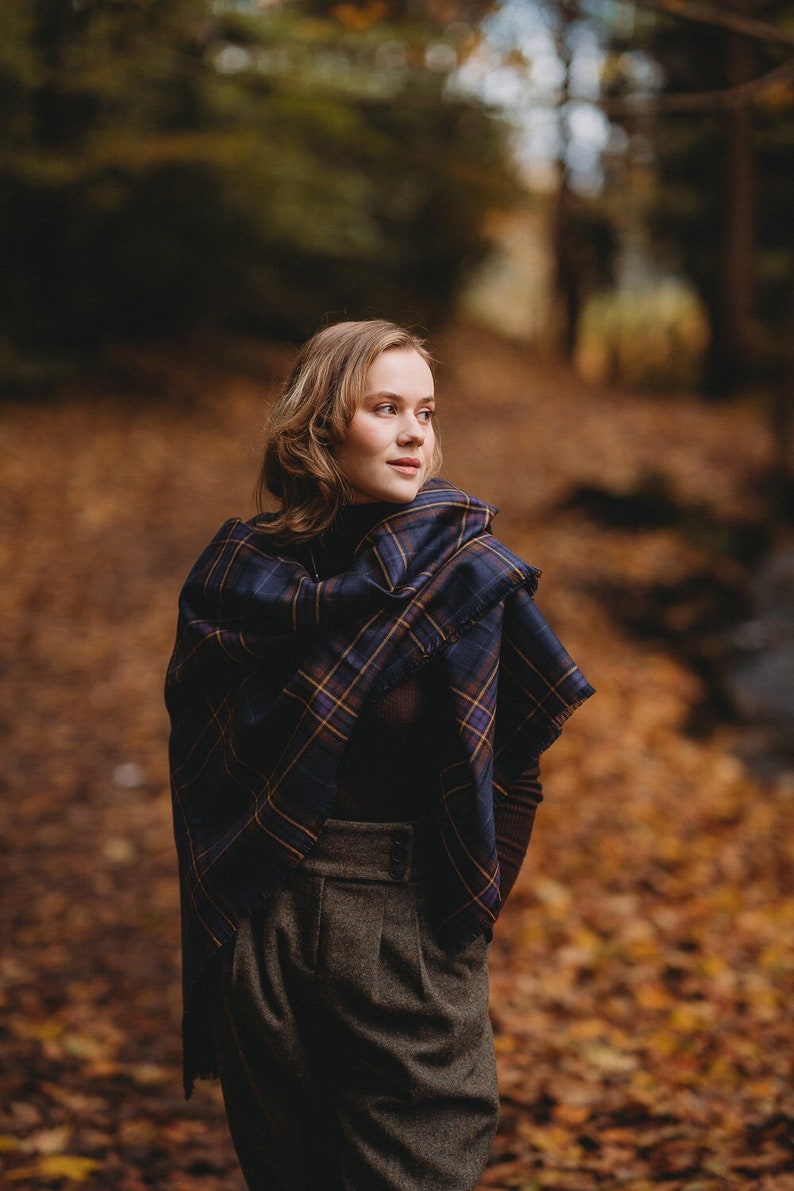 May include: A woman wearing a dark blue and brown plaid scarf with a brown turtleneck sweater and brown pants. She is standing in a forest with fallen leaves on the ground.