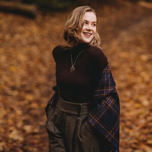 May include: A woman wearing a brown turtleneck sweater with a plaid scarf draped over her shoulders. She is wearing gray high-waisted pants and a gold necklace with a pendant.