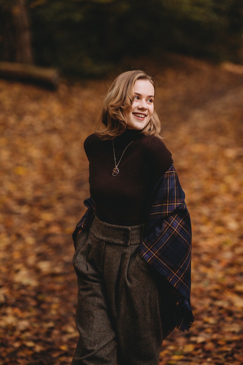 May include: A woman wearing a brown turtleneck sweater with a plaid scarf draped over her shoulders. She is wearing gray high-waisted pants and a gold necklace with a pendant.