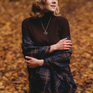 May include: A woman wearing a brown turtleneck sweater and a blue and gold plaid scarf. She is standing in a forest with fallen leaves on the ground. She is wearing a silver necklace with a round pendant.