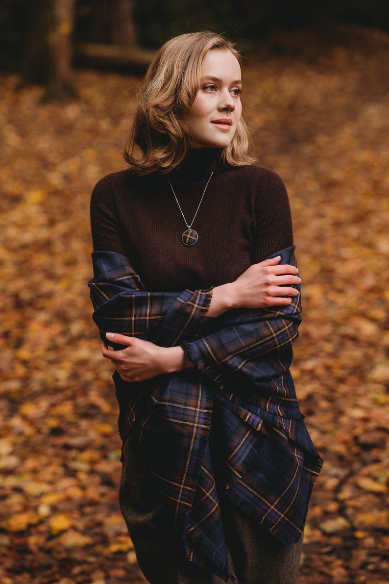 May include: A woman wearing a brown turtleneck sweater and a blue and gold plaid scarf. She is standing in a forest with fallen leaves on the ground. She is wearing a silver necklace with a round pendant.