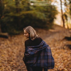 May include: A woman wearing a brown turtleneck sweater and a blue and brown plaid scarf walks through a forest path covered in fallen leaves.