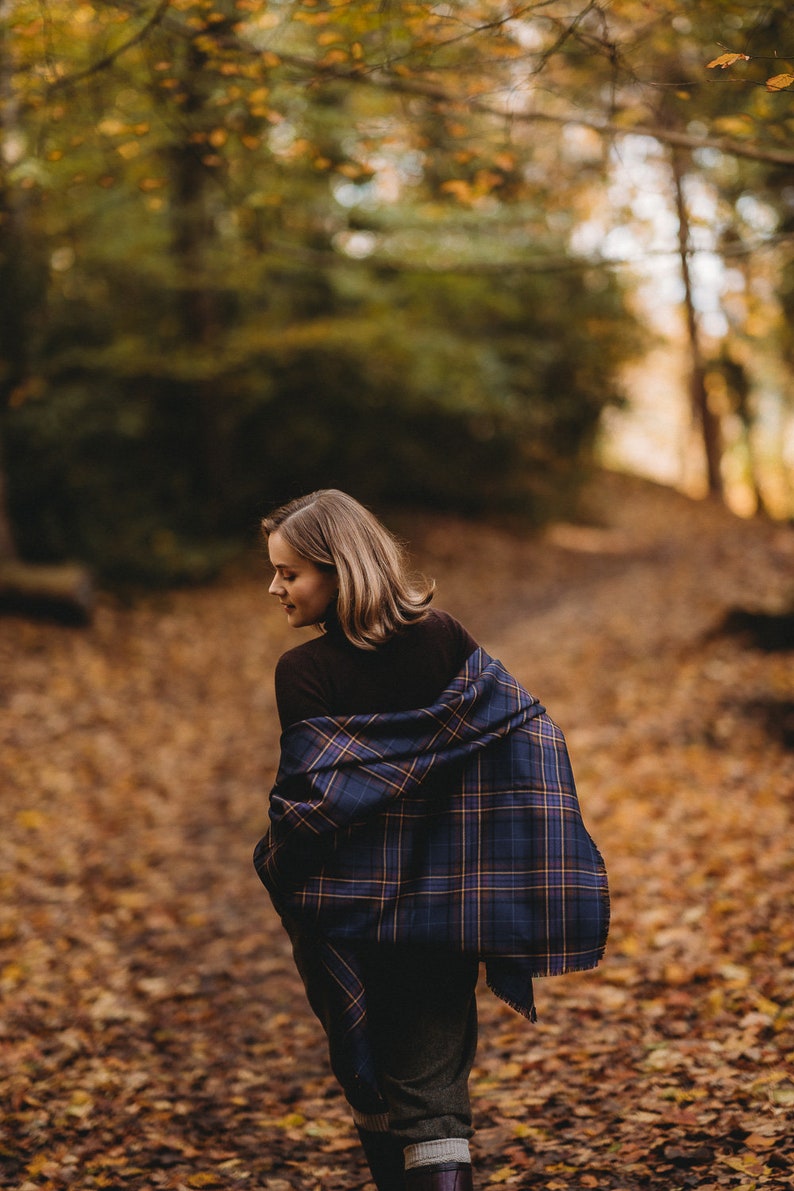May include: A woman wearing a brown turtleneck sweater and a blue and brown plaid scarf walks through a forest path covered in fallen leaves.