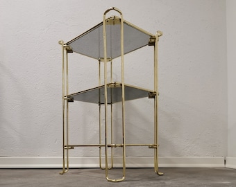 May include: A gold metal three-tiered shelf unit with mirrored glass shelves. The shelves are square and the unit has a simple, geometric design.