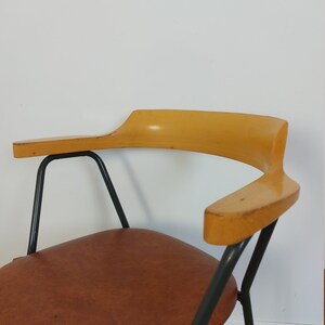 Op de afbeelding: Een vintage stoel met een bruine leren zitting en gebogen houten armleuningen. De stoel heeft een zwart metalen frame en een minimalistisch ontwerp, perfect voor een woonkamer of kantoor.