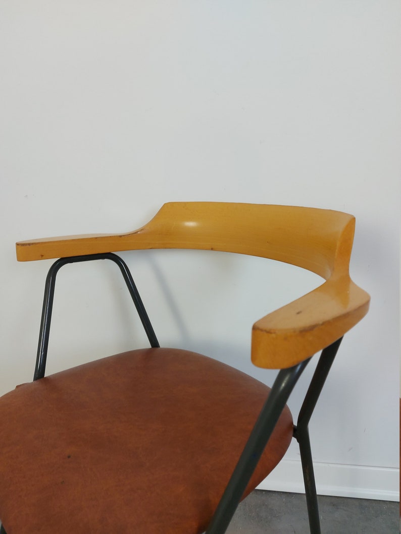 Op de afbeelding: Een vintage stoel met een bruine leren zitting en gebogen houten armleuningen. De stoel heeft een zwart metalen frame en een minimalistisch ontwerp, perfect voor een woonkamer of kantoor.