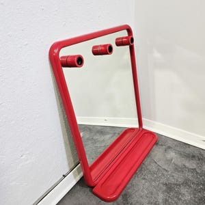 Op de afbeelding: Een rode, rechthoekige spiegel met een afgerond frame en een dienbladachtige basis. Drie cilindrische, rode accenten zijn aan de bovenkant van de spiegel bevestigd. De spiegel leunt tegen een witte muur.