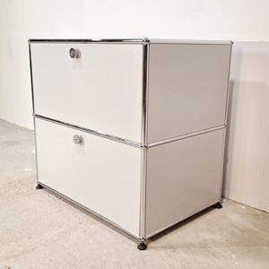 May include: A white two-drawer cabinet with chrome trim and small, round silver knobs. The cabinet is made of white panels and sits on small black feet. The design is minimalist and modern, suitable for storage.