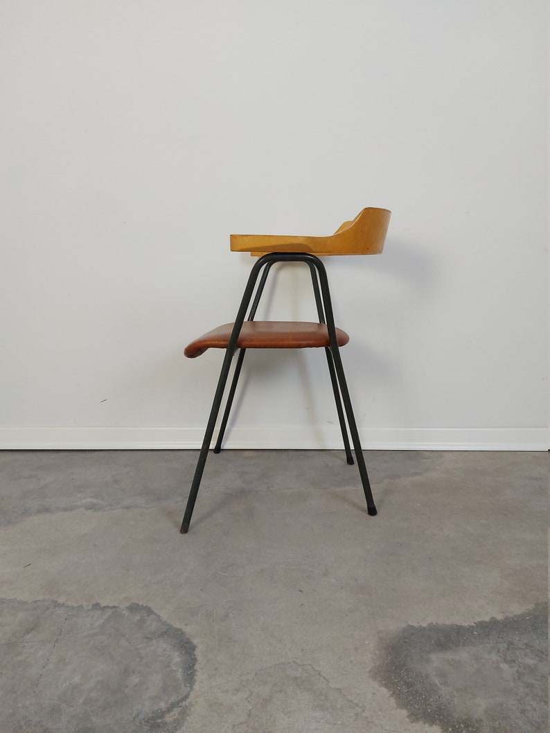 Op de afbeelding: Een vintage stoel met een houten rugleuning en zitting, ondersteund door zwarte metalen poten. De zitting is bruin en de rugleuning is licht van kleur. De stoel heeft een uniek ontwerp, met een gebogen rugleuning en armleuning.