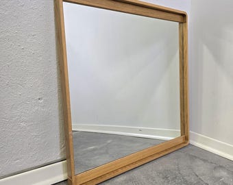 May include: Square wooden framed mirror. The frame is made of light-colored wood with rounded corners. The mirror is leaning against a white wall and a gray floor.
