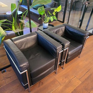 Könnte beinhalten: Zwei schwarze Leder-Sessel mit verchromten Gestellen, die auf einem Holzboden stehen. Die Sessel stehen in einem Raum mit einem großen Fenster und einer grünen Pflanze im Hintergrund.