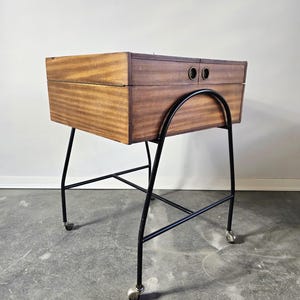 May include: A mid-century modern wooden cabinet with two drawers, mounted on a black metal frame with wheels. The cabinet has a warm brown finish and two circular drawer pulls. The metal frame has a curved design.