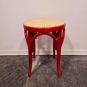 Könnte beinhalten: Ein roter Holzhocker mit einem geflochtenen Korbsitz. Der Hocker hat ein schlichtes, klassisches Design mit geschwungenen Beinen.