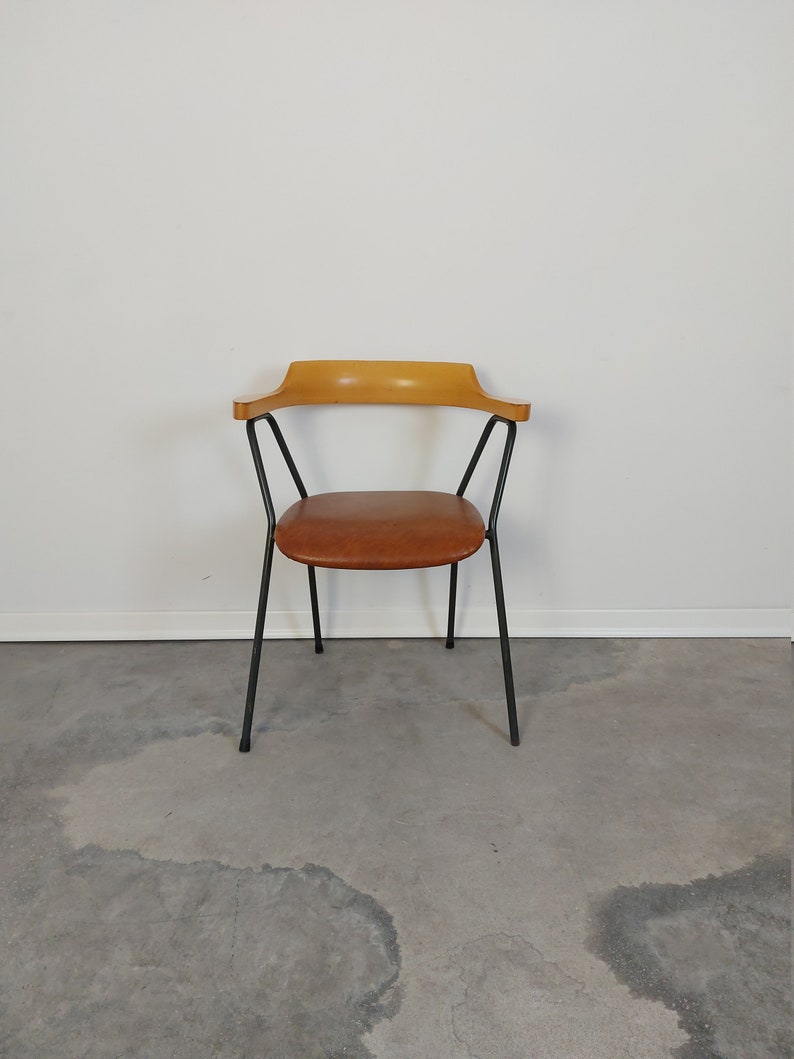 Op de afbeelding: Een stoel in mid-century stijl met een gebogen houten rugleuning en armleuningen. De zitting is bruin en de poten zijn van zwart metaal. De stoel staat tegen een witte muur en een grijze betonnen vloer.
