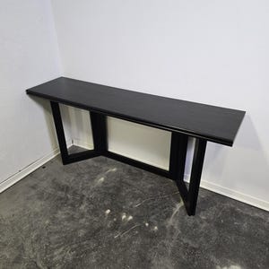 May include: A long, black console table with a rectangular top and angled legs. The table is made of wood and has a minimalist design. The table is approximately 152 cm long.