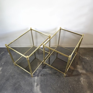 May include: Two square side tables with brass frames and glass tops. The tables are placed together, demonstrating a minimalist design. The brass frames have rounded details at the corners and base. Suitable for a living room or bedroom. Each table is approximately 50cm x 50cm.