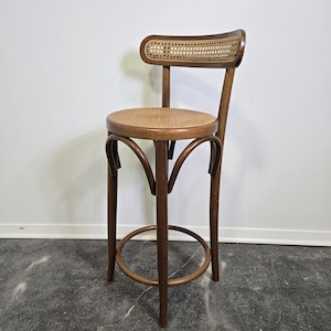 May include: A tall, wooden bar stool with a woven seat and backrest. The stool has a curved back and a circular footrest. The wood is a warm brown color, and the woven elements are a lighter tan. The stool is designed for a bar or counter.