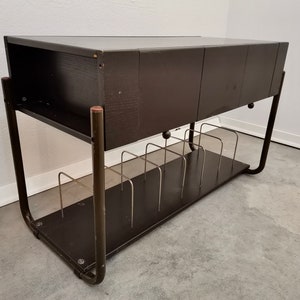 Può includere: Un supporto per giradischi in metallo nero con un piano in legno e uno scaffale per riporre i dischi. Il supporto ha un cassetto per riporre altri oggetti.