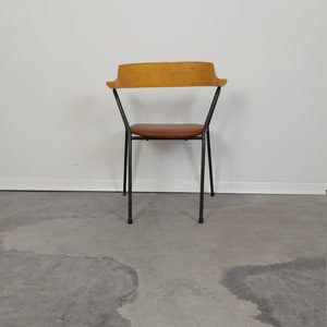 Op de afbeelding: Een stoel in mid-century modern stijl met een lichtbruine houten rugleuning en een bruine leren zitting. De stoel heeft een zwart metalen frame en vier poten. De stoel staat tegen een witte muur en een grijze betonnen vloer.