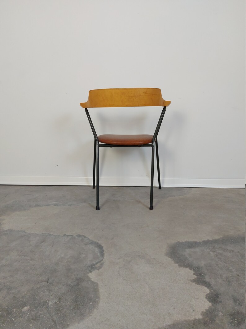 Op de afbeelding: Een stoel in mid-century modern stijl met een lichtbruine houten rugleuning en een bruine leren zitting. De stoel heeft een zwart metalen frame en vier poten. De stoel staat tegen een witte muur en een grijze betonnen vloer.