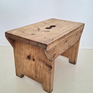 May include: A rustic, wooden step stool with a rectangular seat and four sturdy legs. The stool is made of light-colored wood with visible grain and a natural finish. A hand-hold is cut into the seat. The design is simple and functional.