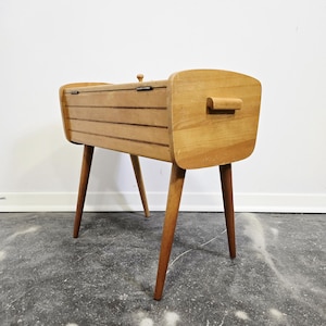 Op de afbeelding: Een vintage houten naaidoos met een scharnierend deksel en een handvat. De doos is lichtbruin en heeft een rechthoekige vorm met vier taps toelopende poten. Het deksel heeft een kleine houten knop. Ontworpen voor het opbergen van naaigerei.