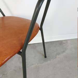 Op de afbeelding: Een stoel in mid-century stijl met een gebogen houten rugleuning en een bruine zitting. De stoel heeft een donkergrijs metalen frame en poten. De zitting lijkt van leer of een leerachtig materiaal te zijn. De stoel staat tegen een witte muur en een grijze betonnen vloer.