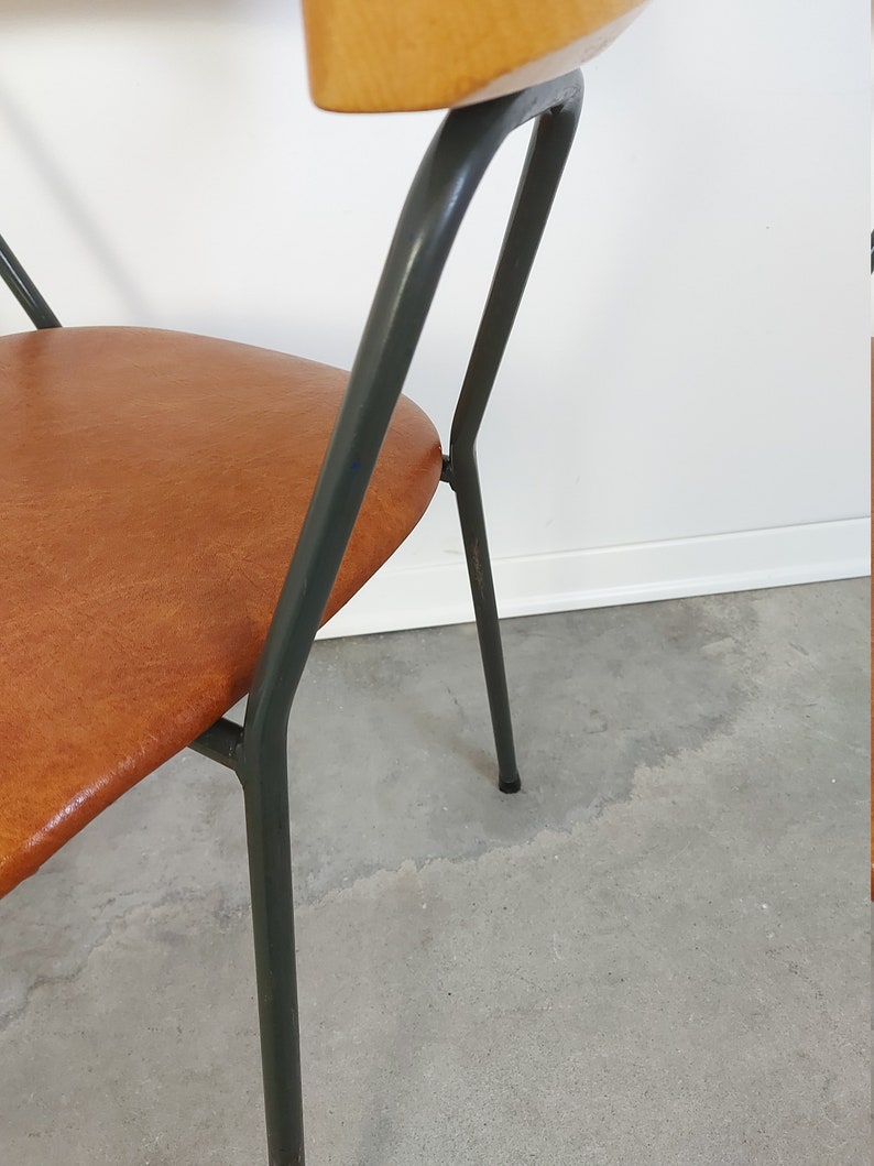 Op de afbeelding: Een stoel in mid-century stijl met een gebogen houten rugleuning en een bruine zitting. De stoel heeft een donkergrijs metalen frame en poten. De zitting lijkt van leer of een leerachtig materiaal te zijn. De stoel staat tegen een witte muur en een grijze betonnen vloer.