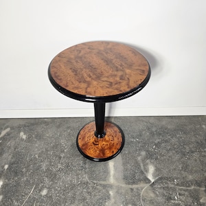 May include: A small, round side table with a dark brown, burl wood top and base. The table has a black, cylindrical stem and a black trim around the top and base. The table is approximately 61 cm tall.