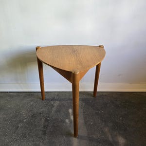 Peut inclure: Petite table d'appoint triangulaire en bois clair, avec trois pieds. Le plateau affiche un veinage visible. Les pieds cylindriques et élancés sont également en bois clair. Le tout est posé devant un mur blanc et un sol gris foncé.