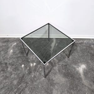 Pode incluir: Uma mesa de centro quadrada com tampo de vidro e estrutura de metal cromado. A mesa tem quatro pernas e está sobre um piso de concreto cinza.
