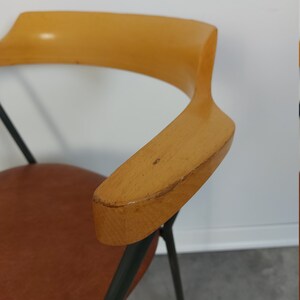 Op de afbeelding: Mid-century moderne stoel met een lichtbruine houten armleuning en een bruine leren zitting. De stoel heeft zwarte metalen poten en een gebogen armleuningontwerp. De achtergrond is een witte muur en een grijze vloer.