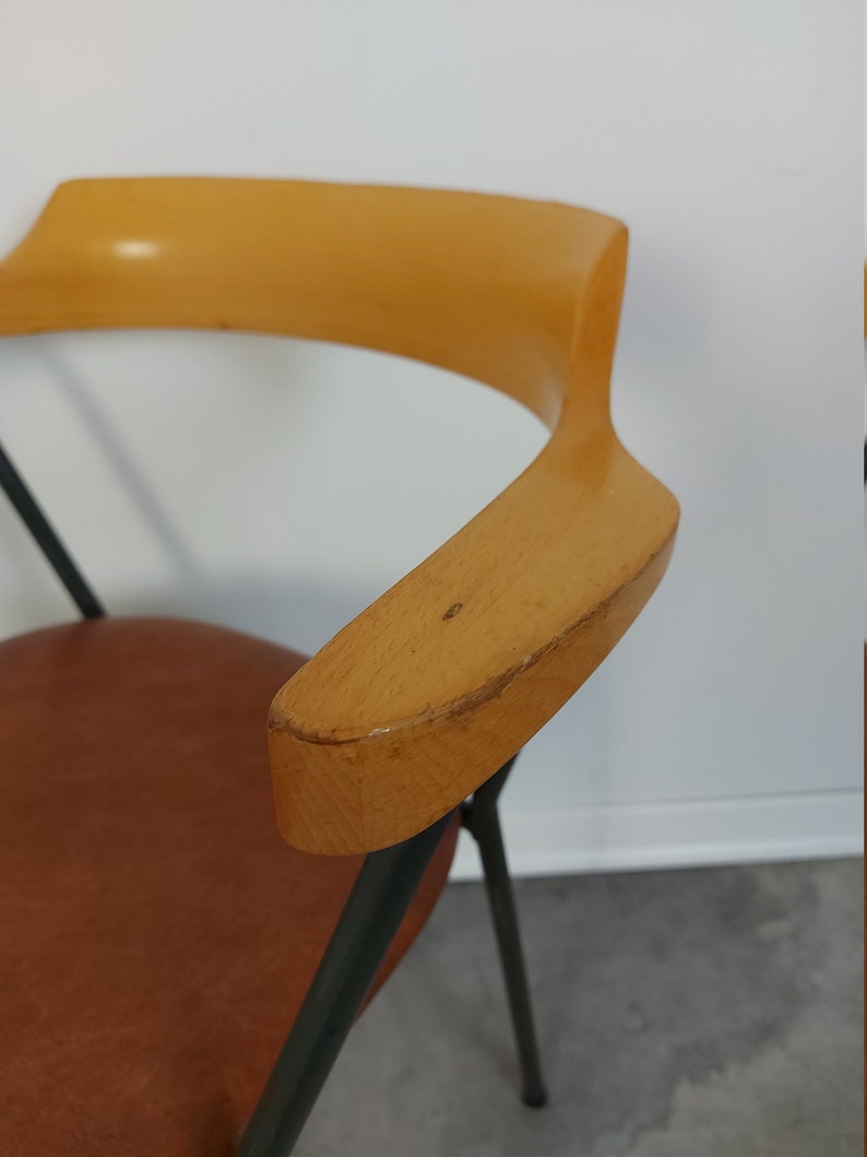 Op de afbeelding: Mid-century moderne stoel met een lichtbruine houten armleuning en een bruine leren zitting. De stoel heeft zwarte metalen poten en een gebogen armleuningontwerp. De achtergrond is een witte muur en een grijze vloer.