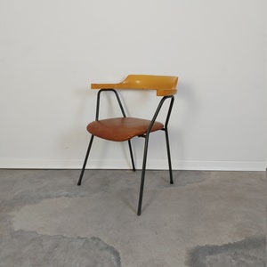 Op de afbeelding: Mid-century moderne stoel met een bruine leren zitting en een gebogen houten armleuning. De stoel heeft een zwart metalen frame en poten. Het ontwerp is minimalistisch, tegen een witte muur en een grijze betonnen vloer.
