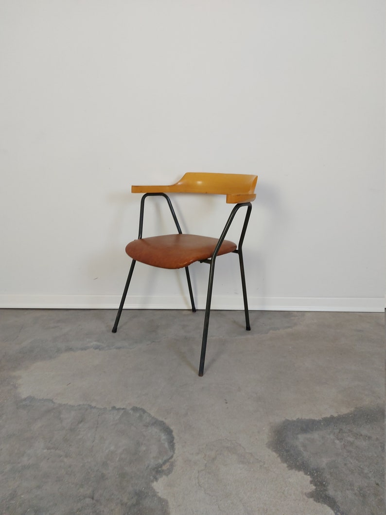 Op de afbeelding: Mid-century moderne stoel met een bruine leren zitting en een gebogen houten armleuning. De stoel heeft een zwart metalen frame en poten. Het ontwerp is minimalistisch, tegen een witte muur en een grijze betonnen vloer.