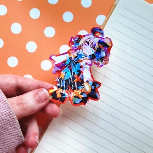 May include: A colorful, iridescent sticker featuring an anime-style character with long blonde hair, wearing a black and white outfit with a red and yellow design. The sticker is on a white and orange polka dot background.