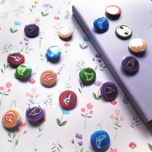 May include: A collection of colorful, round buttons with various symbols and designs. The buttons are arranged on a floral patterned surface next to a purple notebook. The buttons feature a variety of colors including red, green, purple, and blue.