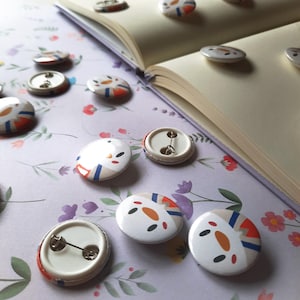 May include: A collection of white, round buttons with colorful abstract designs in orange, blue, and black. Some buttons are scattered on a floral patterned surface near an open book.