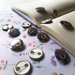 May include: A collection of round, black and gray button pins with a gold "C" design, scattered on a floral patterned surface next to an open book. The pins have a metal pin back.