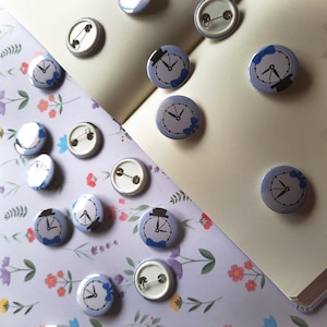 May include: A collection of round, light blue pinback buttons with clock faces and black hands. Some buttons feature a top hat and bow tie design. The buttons are scattered on a floral patterned surface and a cream-colored notebook.