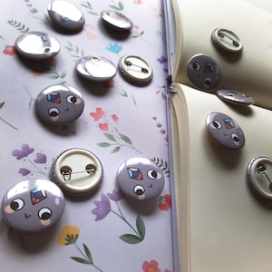 May include: A collection of round, gray enamel pins with a cartoon face design and colorful gem details. Some pins are face-up, while others show the pin back. The pins are arranged on a floral patterned surface and an open book.