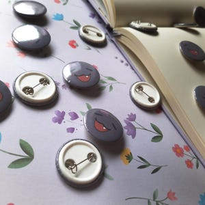 May include: A collection of round, purple enamel pins with a smiling face design. The pins are scattered on a floral patterned surface, with some pins open on a book. The pins have a metal pin back.