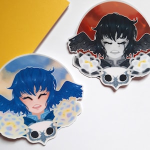 May include: Two round stickers featuring illustrations of a person with blue and black wings. The person on the left sticker has blue hair and is smiling. The person on the right sticker has black hair and is crying. Both stickers have a white flower design and a gray skull-like design.