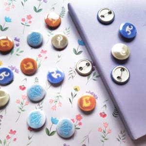 May include: A collection of colorful, round buttons and pins with various symbols and designs. The buttons are orange, blue, and white, with intricate graphics. Some pins have arrow designs. The items are arranged on a floral patterned background next to a purple notebook.