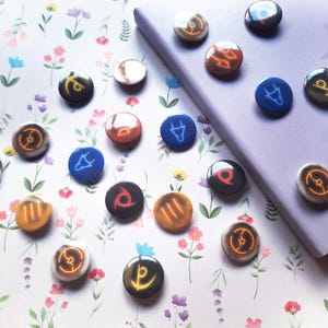 Può includere: Una collezione di magneti rotondi e lucidi con vari simboli arancioni, blu e bianchi. I magneti sono disposti su una superficie a motivi floreali e un taccuino viola. I simboli sono stilizzati e sembrano essere a tema fantasy.