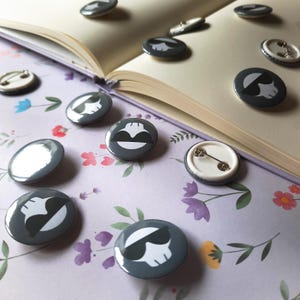 May include: A collection of gray button pins with white and black graphic designs, including a skull with sunglasses and a smiling face. The pins are scattered on a floral patterned surface and an open book.