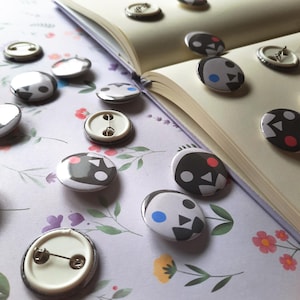 May include: A collection of round, button-style pins with a black and white graphic design, scattered on a floral patterned surface. Some pins are face-up, revealing the pin backings. An open book is in the background.