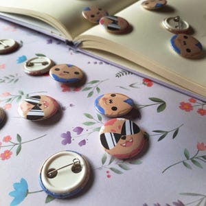 May include: A collection of round pinback buttons with cartoon character designs. The buttons feature various faces with sunglasses, stripes, and blue accents. Some buttons are face-up, while others show the pin backing. The buttons are scattered on a floral patterned surface.