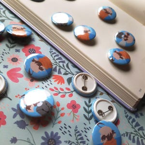 May include: A collection of round, blue pinback buttons with cartoon character illustrations. Some buttons show the character's face, while others show the back with a pin. The buttons are arranged on a floral patterned surface and a book.