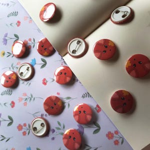 May include: A collection of red, round pinback buttons with a cartoon animal design. The buttons are scattered on a floral patterned paper and a blank notebook page. Each button has a safety pin on the back.